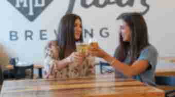 Two women cheers their beers and smile inside the tasting room of Mirror Twin Brewing.