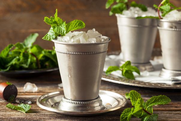 Mint Julep cocktail in an antique, silver cocktail glass sits on a silver platter with fresh sprigs of mint in the drink and on the wooden table.