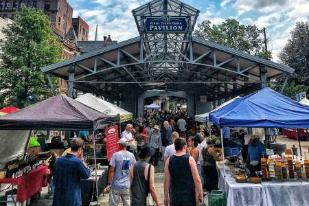 A photo of the Lexington Farmers Market at the Fifth Third Pavilion. People can be seen shopping at different vendor’s tents. The sky above the pavilion is a beautiful blue color with clouds peppered throughout.