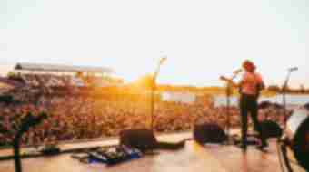 A woman plays guitar on stage for a a huge crowd in a field as the sun sets behind them.