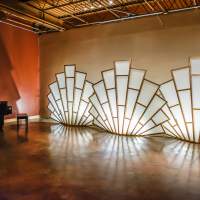 Three large, glowing, white and gold art deco-style installations and a grand piano in a large room with hardwood flooring.